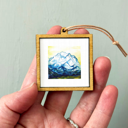 A person's hand holding a small wooden frame containing a framed painting of a mountain landscape.