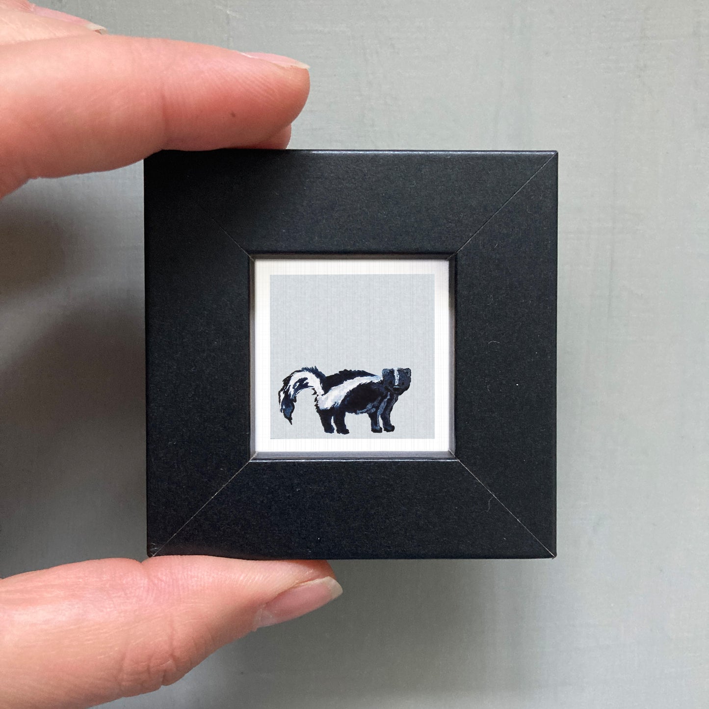 A hand is holding a small black square frame containing a black and white illustration of a skunk.