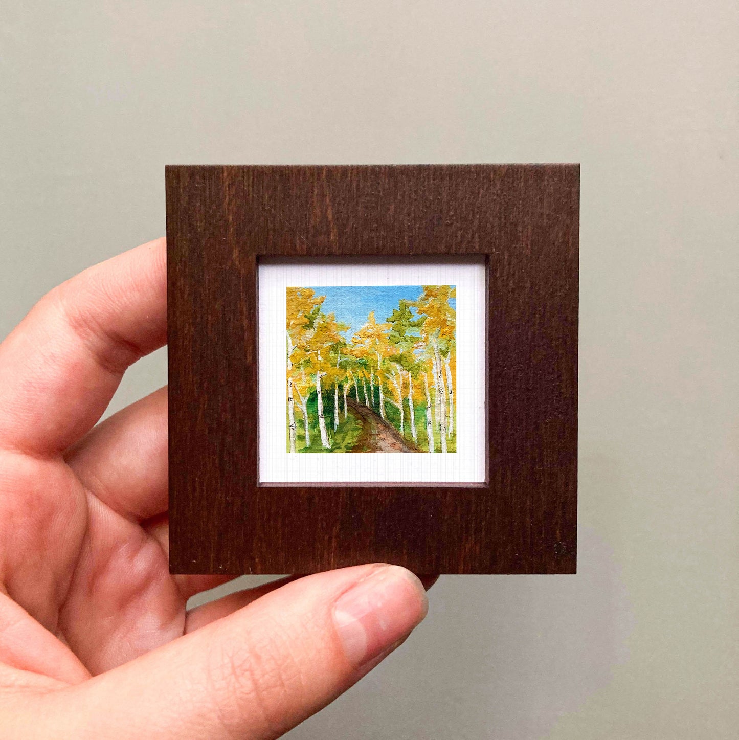 A hand is holding a small, square-shaped wooden frame containing a painting of a forest with tall trees and a path.