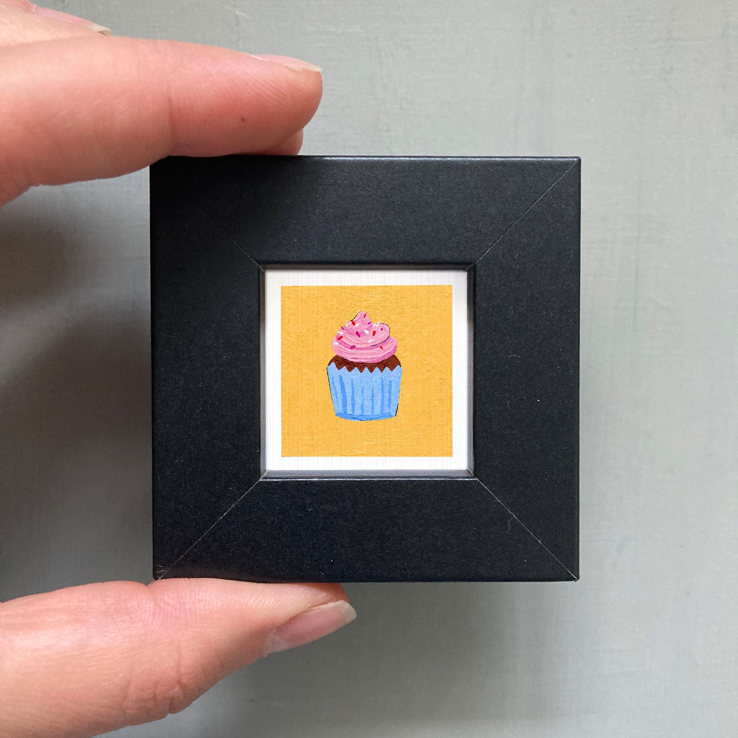 A hand is holding a small black frame containing a framed illustration of a cupcake with pink frosting.