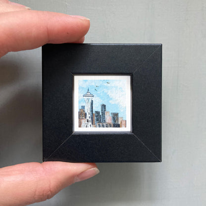 A person's hand holding a small black frame containing a miniature painting of the Seattle skyline.