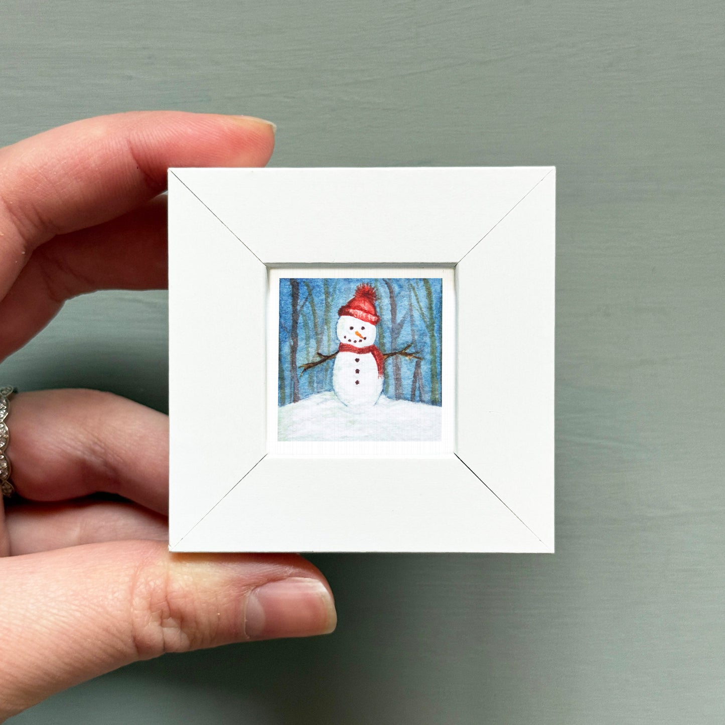A hand is holding a small white square frame containing a painting of a snowman.