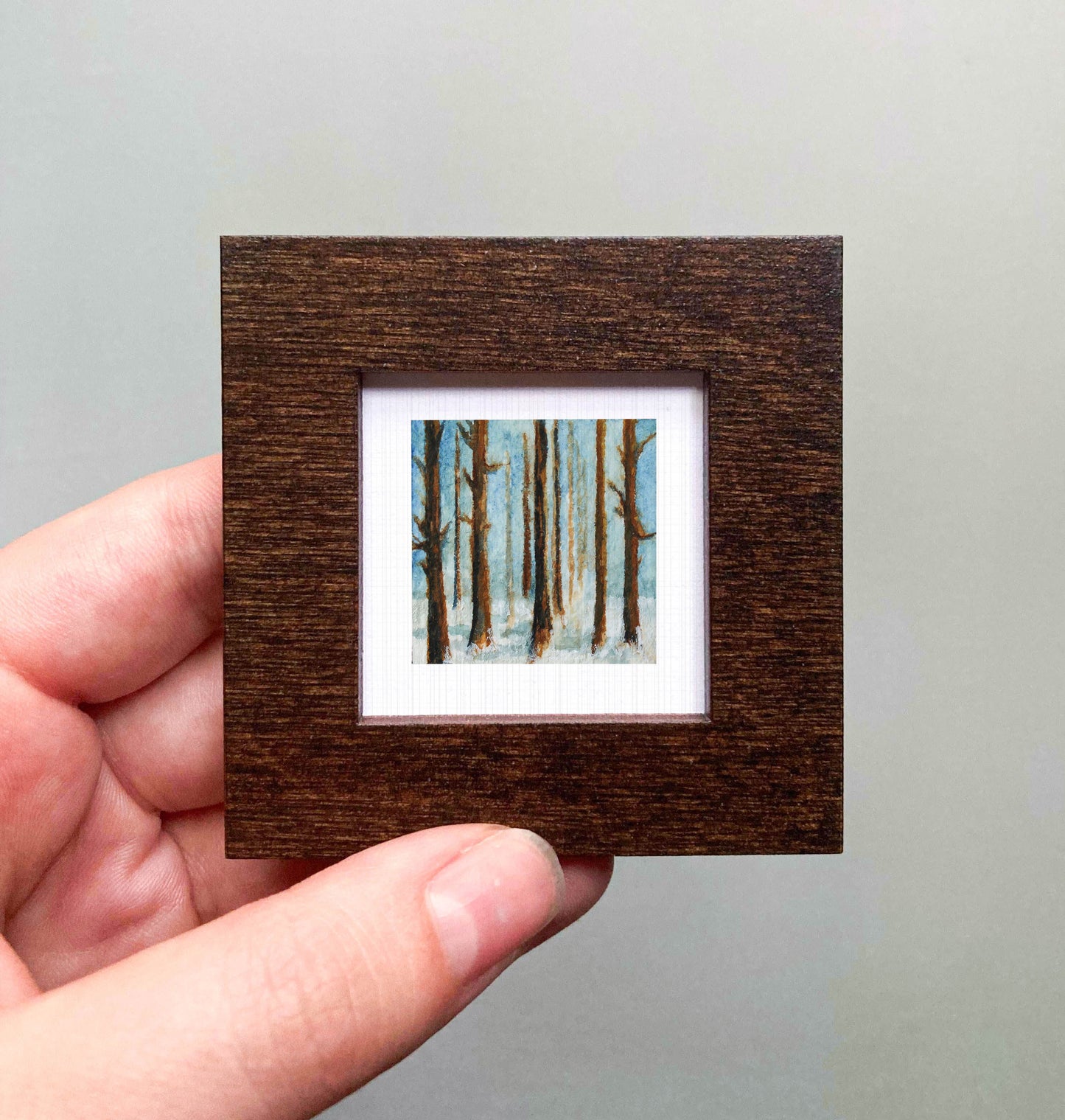 A hand is holding a small, square-shaped wooden frame containing a painting of trees in a forest.