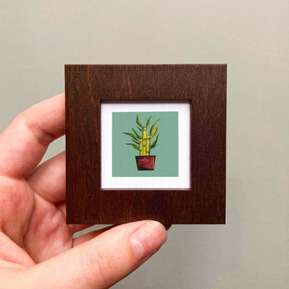 A hand is holding a small wooden frame containing a framed illustration of a potted plant.
