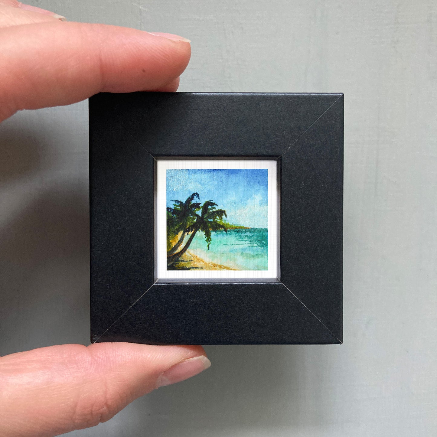 A person's hand holding a small square picture frame containing a painting of a tropical beach scene with palm trees and a blue sky.
