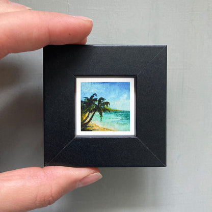 A person's hand holding a small square picture frame containing a painting of a tropical beach scene with palm trees and a blue sky.
