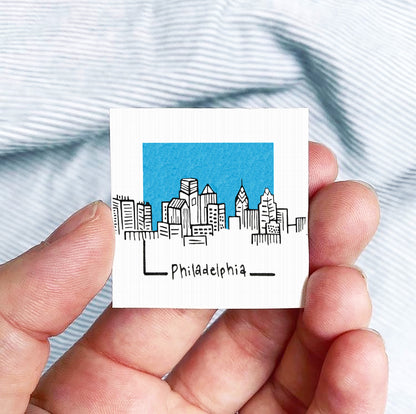 A hand holding a small square-shaped object with a drawing of a city skyline and the word "PhiladELPHIA" written on it.