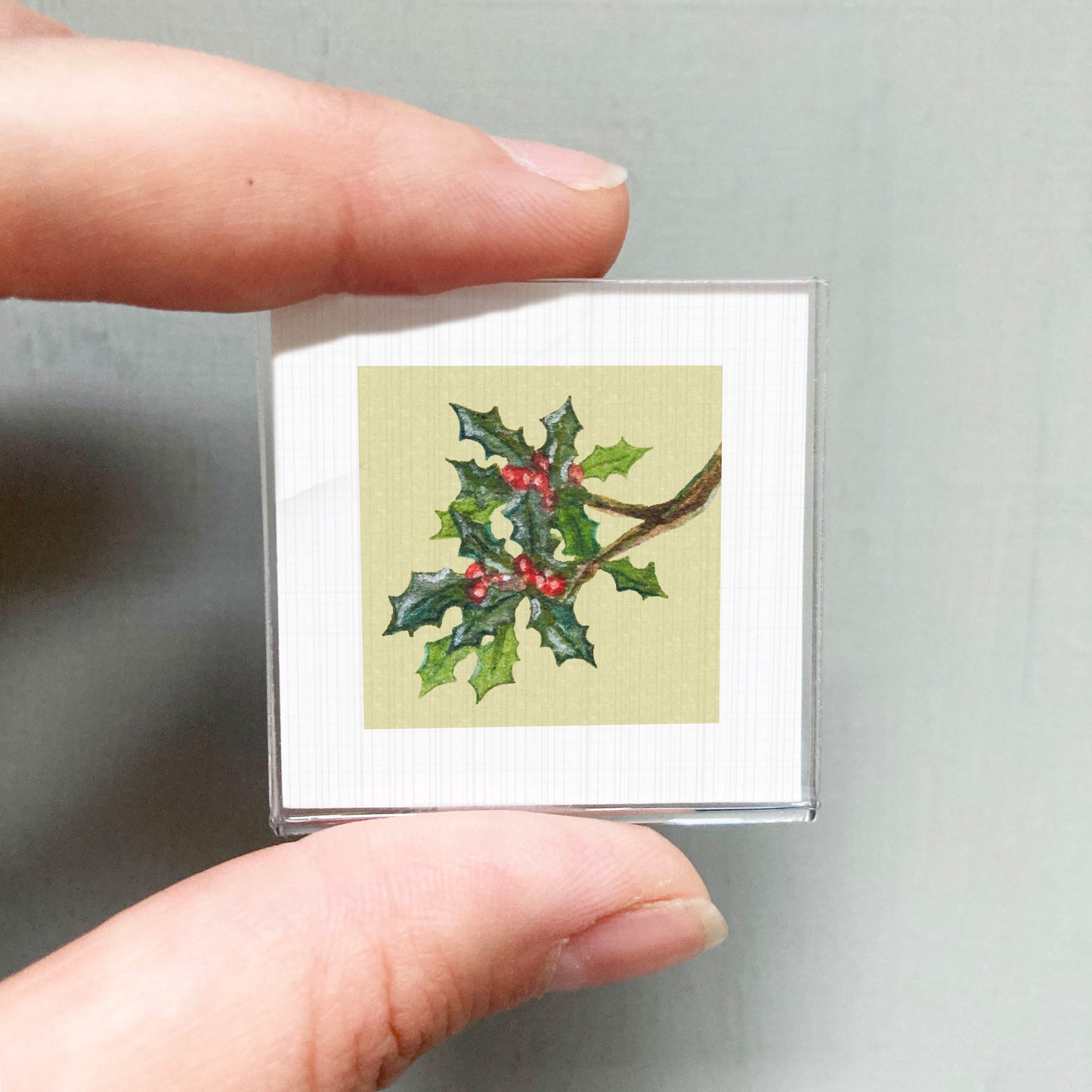 A person's hand holding a small square card with a holly leaf and berries on it.