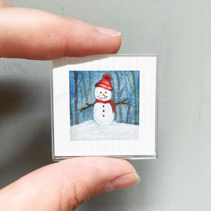 A person's hand is holding a small square picture of a snowman in a snowy forest.