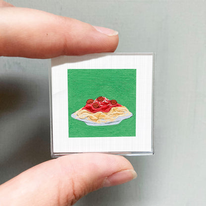A person's hand holding a small square picture depicting a plate of spaghetti with tomato sauce.