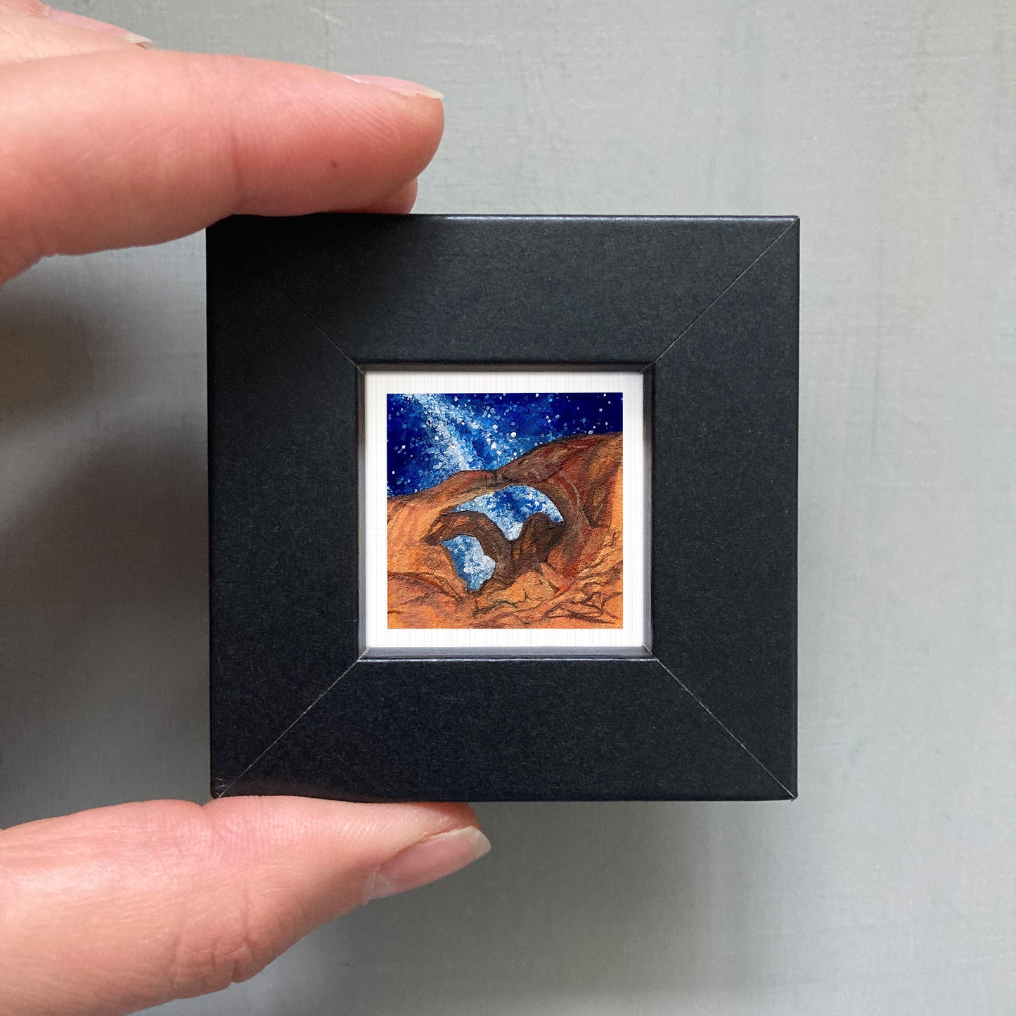 A person's hand holding a small black frame containing a framed painting of a desert landscape with a starry night sky.