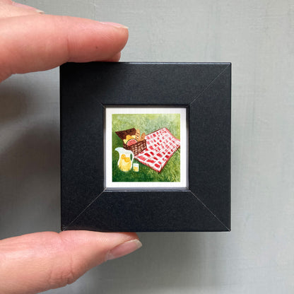 A person's hand holding a small black picture frame containing a framed painting of a picnic scene with a basket, blanket, and food items.