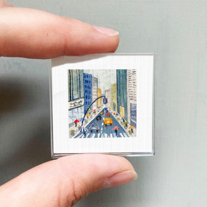 A person's hand is holding a small square picture depicting a city street scene with people walking and cars driving.