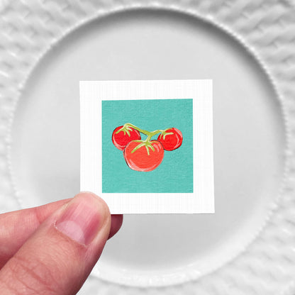 A hand is holding a small square with a vibrant illustration of three red tomatoes against a teal background.