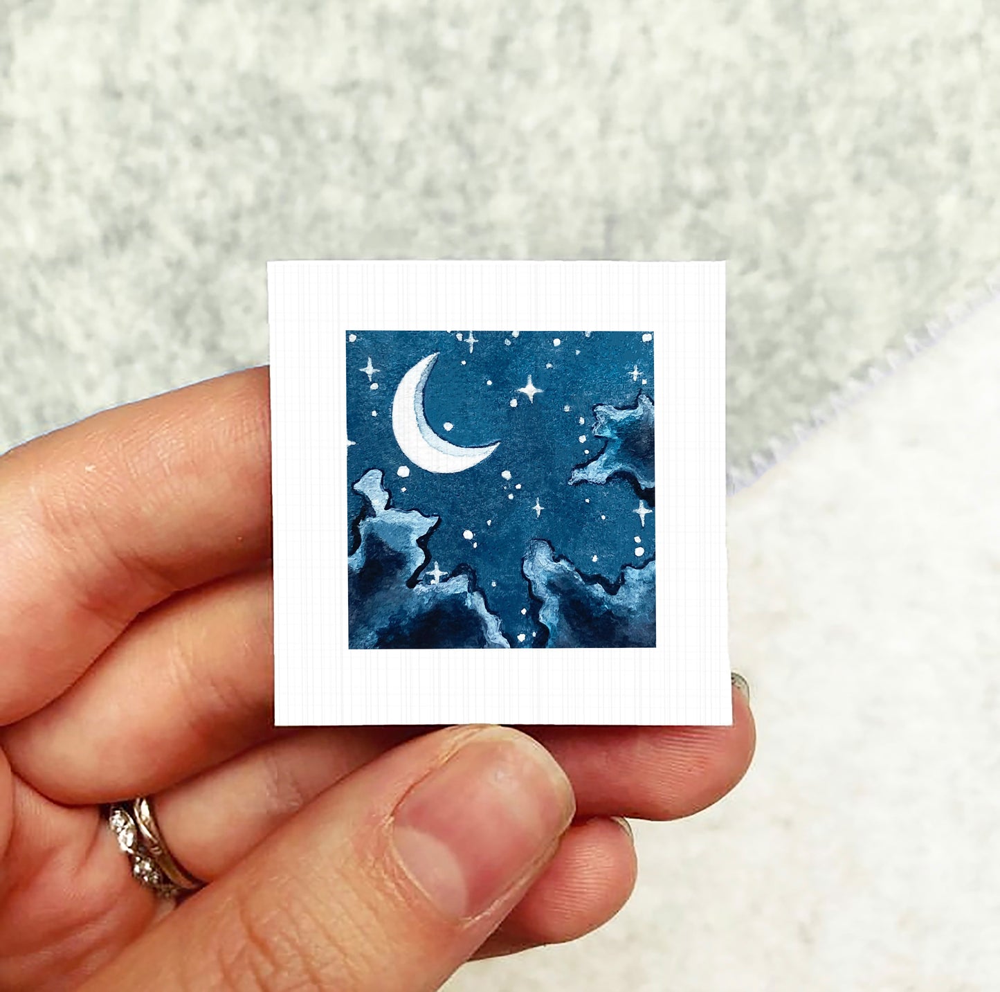 A hand holding a small square-shaped piece of paper with a watercolor painting of a night sky with a crescent moon and stars.