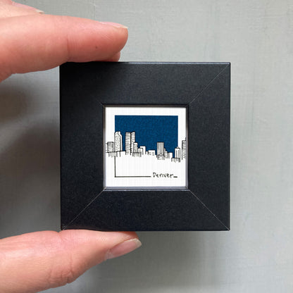 A person's hand holding a small black frame containing a framed illustration of a city skyline.