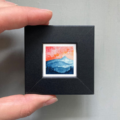 A hand is holding a small, square-shaped black frame containing a watercolor painting of a mountain landscape with a sunset sky.
