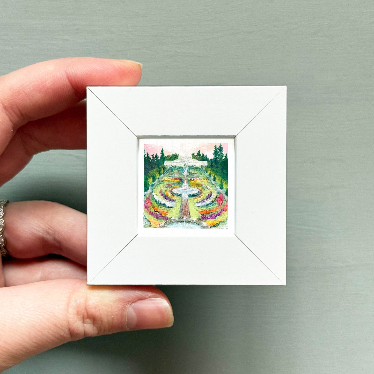 A hand is holding a small white square frame containing a painting of a garden with a fountain and trees.