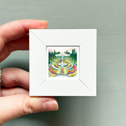 A hand is holding a small white square frame containing a painting of a garden with a fountain and trees.