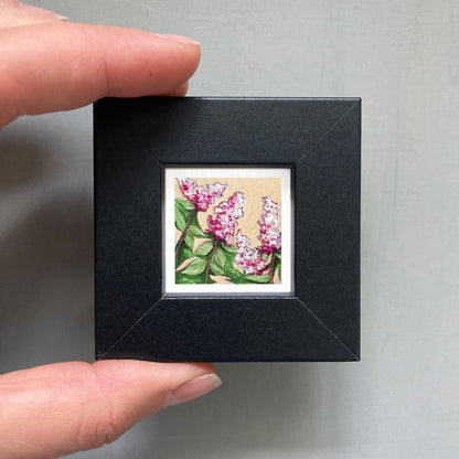 A person's hand holding a small square picture frame containing a painting of pink flowers.