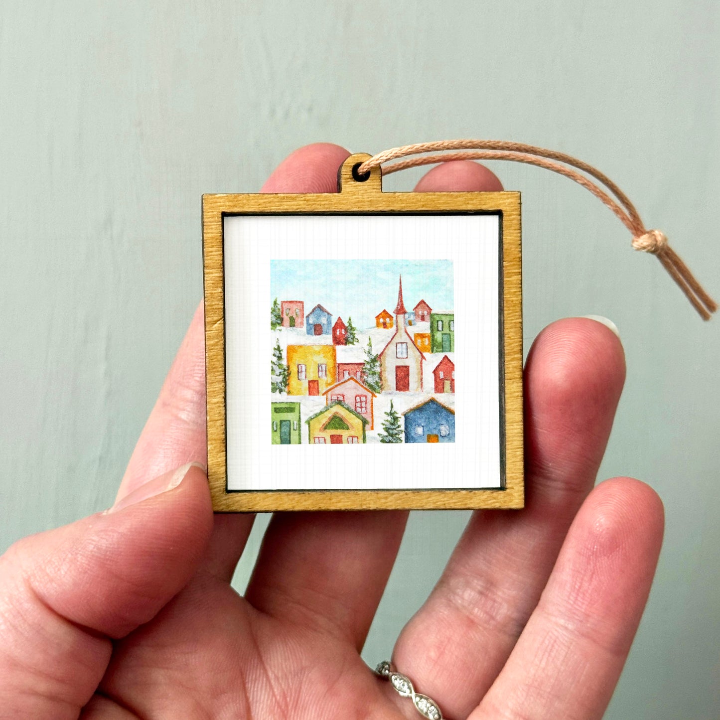 A hand holding a small wooden frame containing a watercolor painting of a colorful village scene.