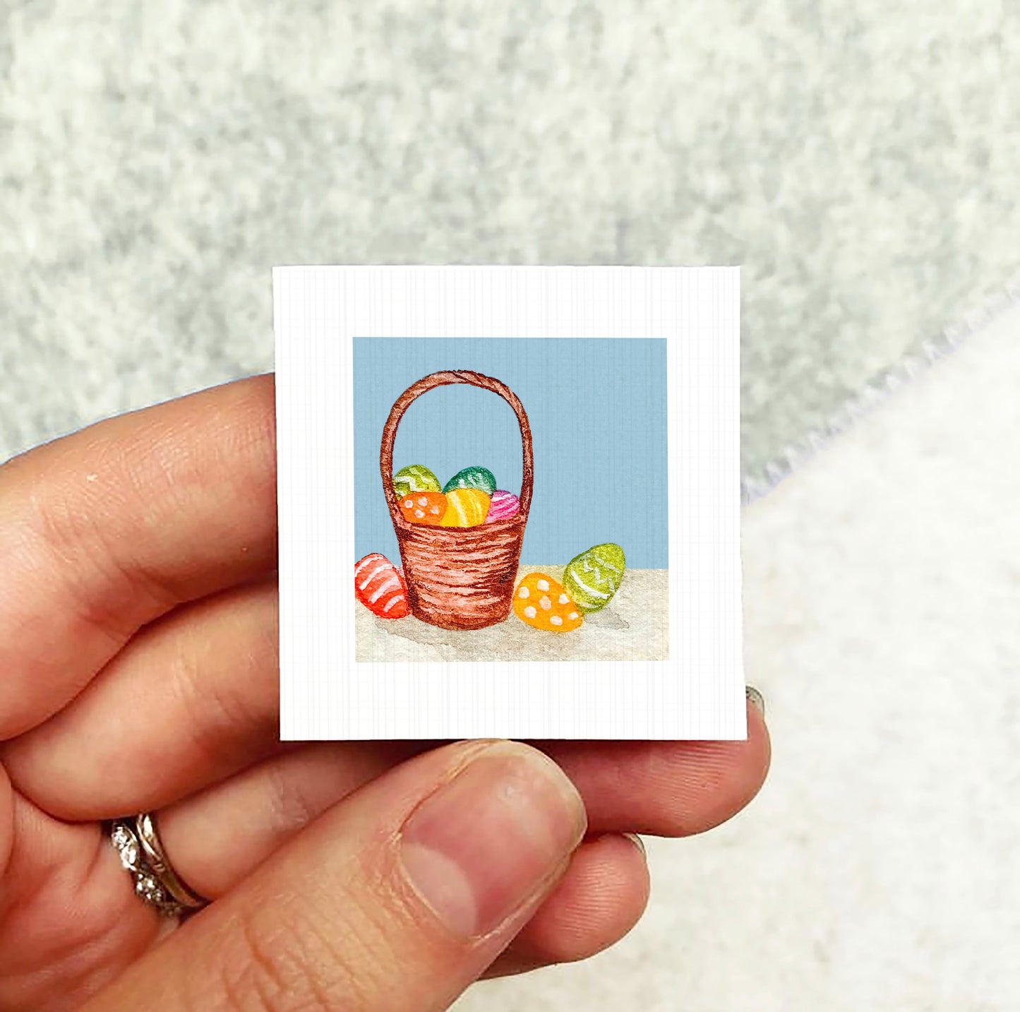 A hand holding a small square picture depicting a basket filled with colorful Easter eggs against a blue background.