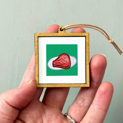 A hand holding a small wooden frame containing a picture of a piece of meat on a white plate against a green background.