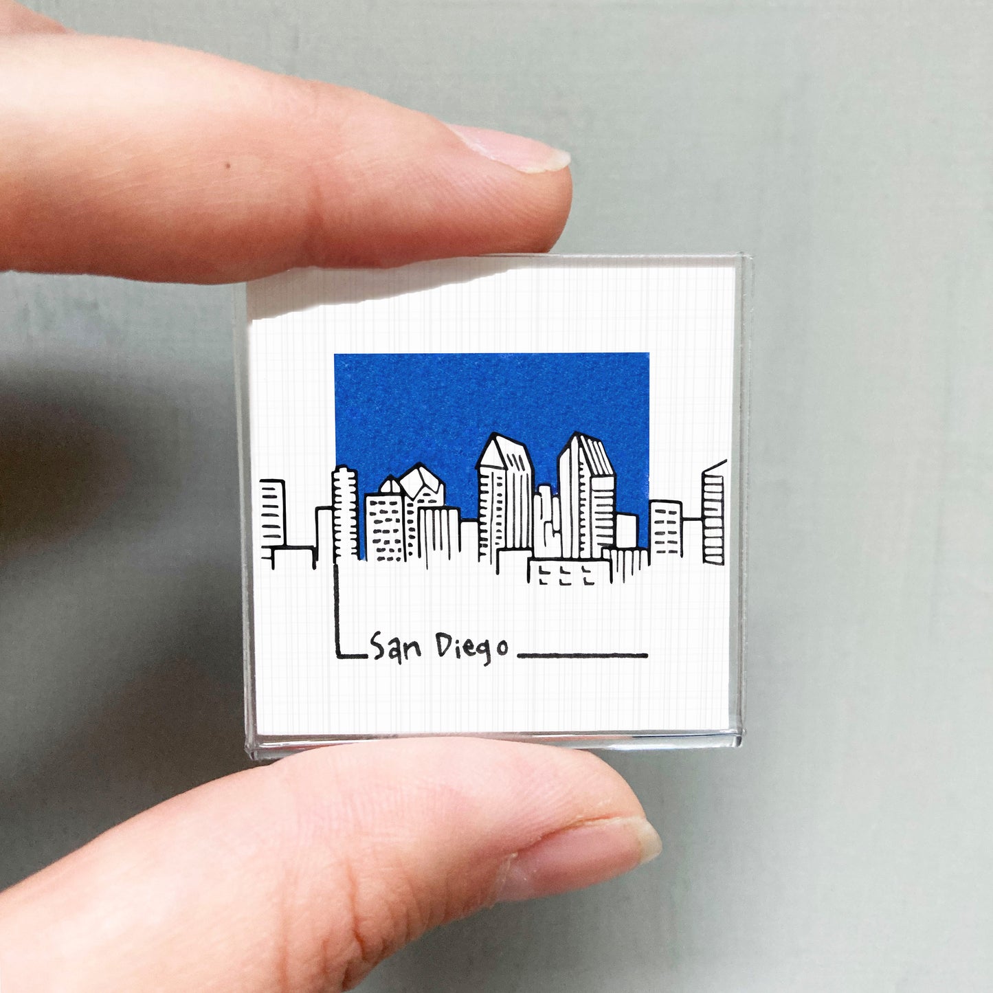 A person's hand holding a small square magnet with a drawing of a city skyline and the text "San Diego" written on it.