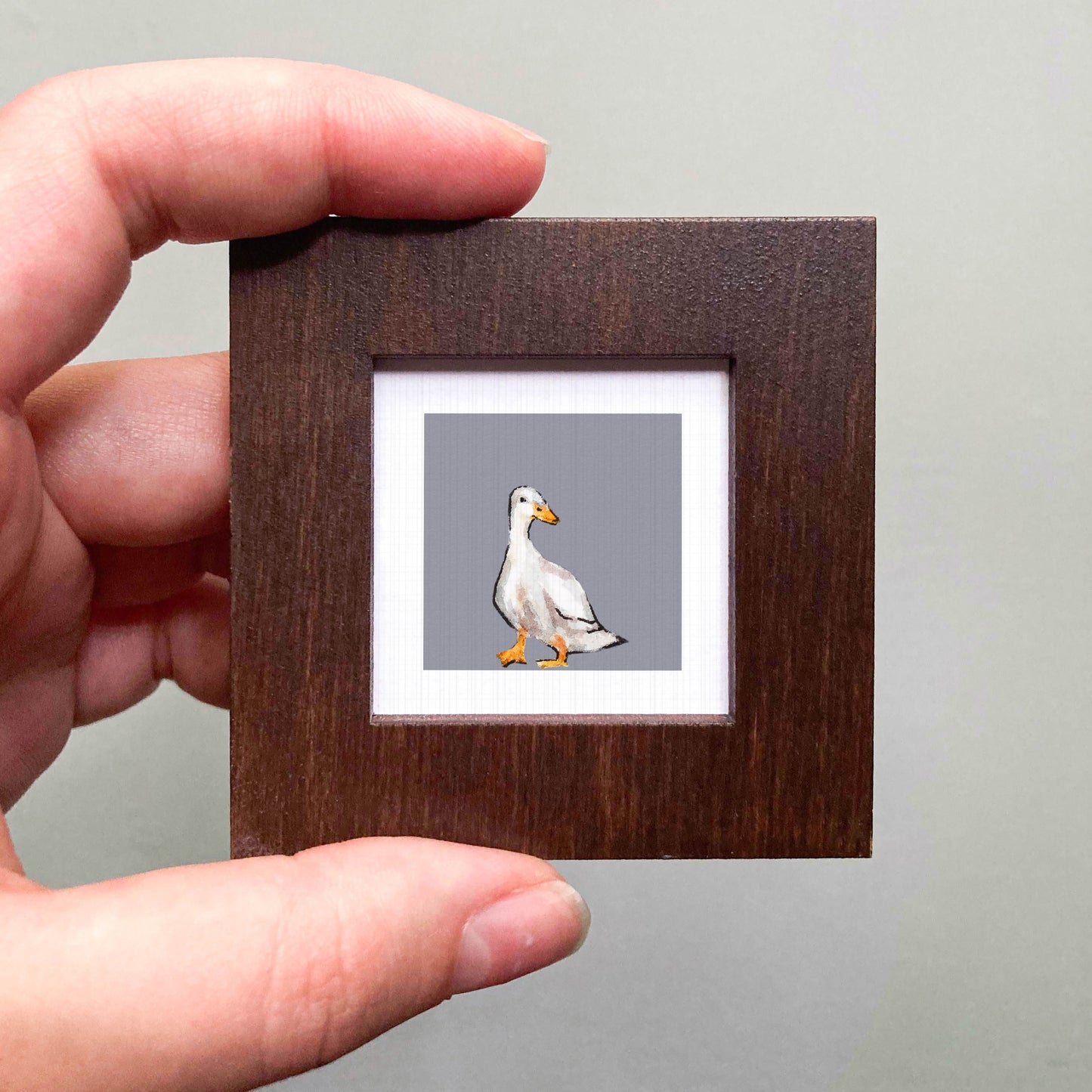 A person's hand is holding a small wooden frame containing a picture of a duck.