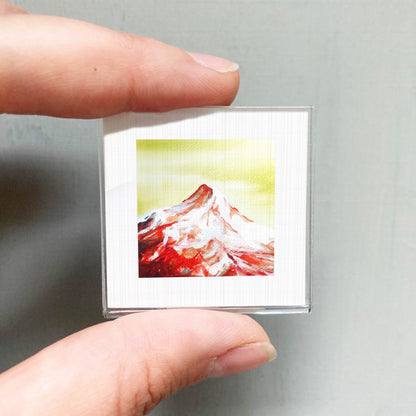 A person's hand holding a small square picture of a mountain with a vibrant red and yellow background.