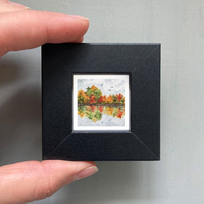 A person's hand holding a small black frame containing a miniature painting of a landscape with trees and a lake.