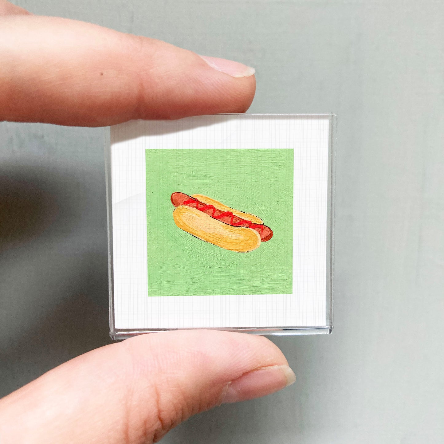 A person's hand holding a small square picture of a hot dog against a green background.