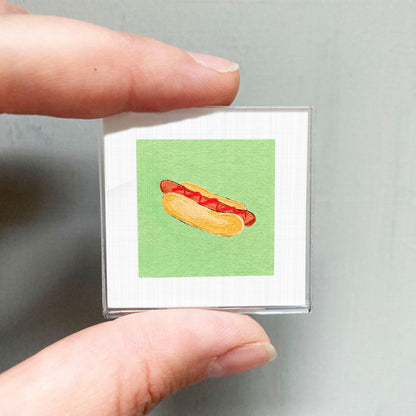 A person's hand holding a small square picture of a hot dog against a green background.