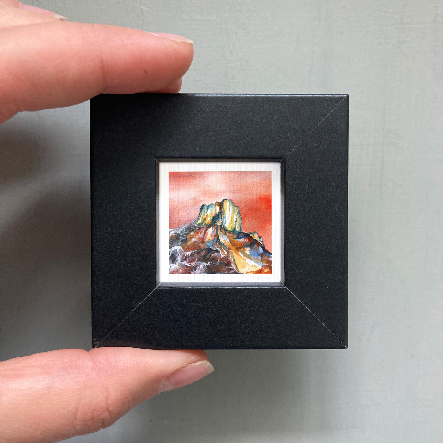 A person's hand holding a small, square-shaped black frame containing a watercolor painting of a mountain landscape.