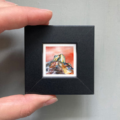 A person's hand holding a small, square-shaped black frame containing a watercolor painting of a mountain landscape.