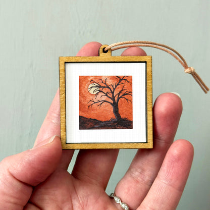 A hand holding a small wooden frame containing a painting of a tree with a full moon in the background.