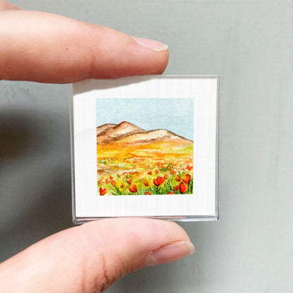 A person's hand holding a small square picture of a field with mountains in the background.