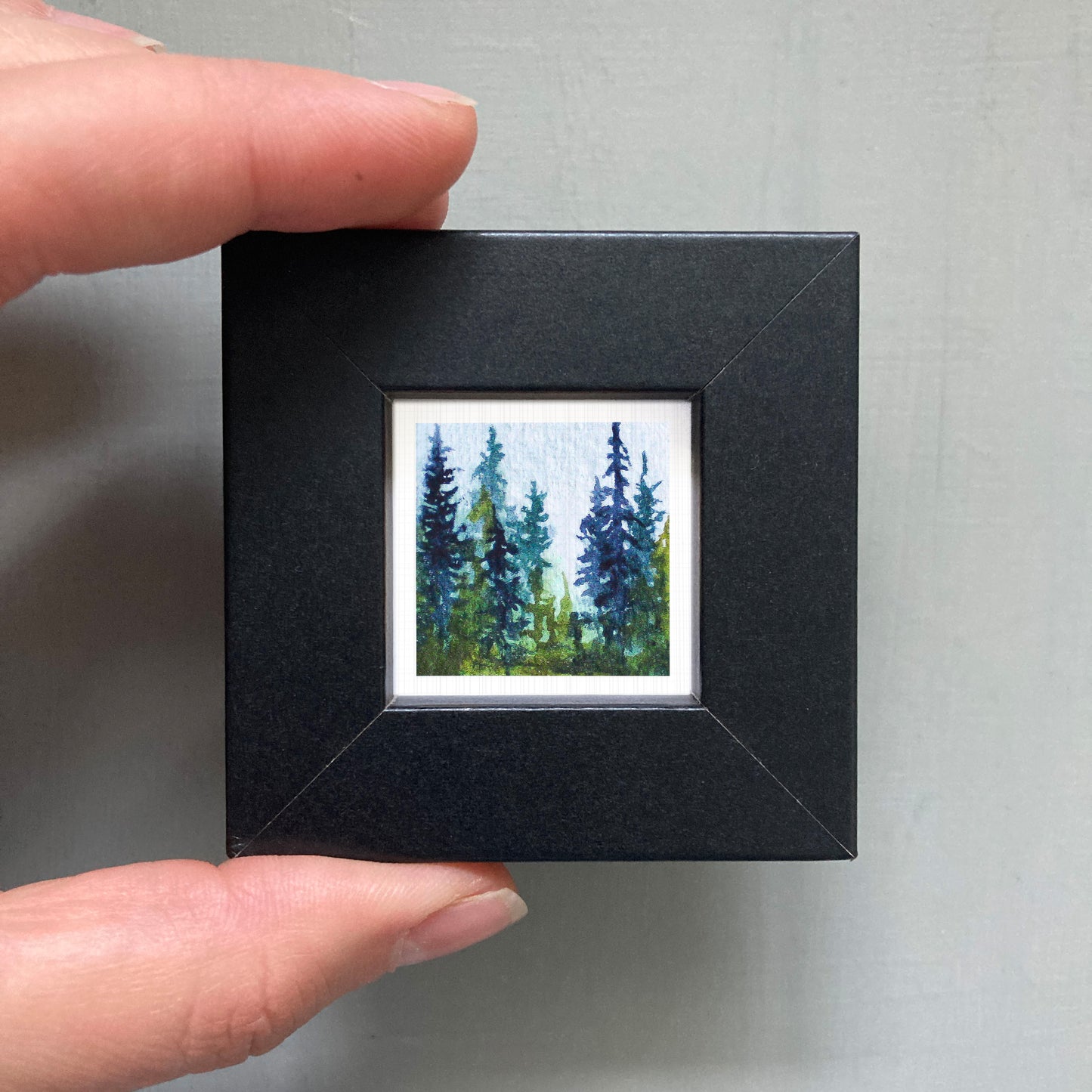 A person's hand holding a small black frame containing a painting of a forest scene with tall trees.