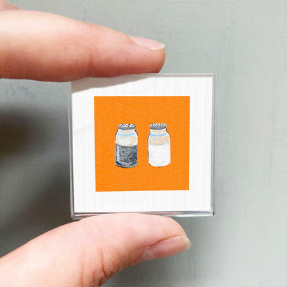 A person's hand holding a small square with an illustration of two bottles of salt and pepper on an orange background.