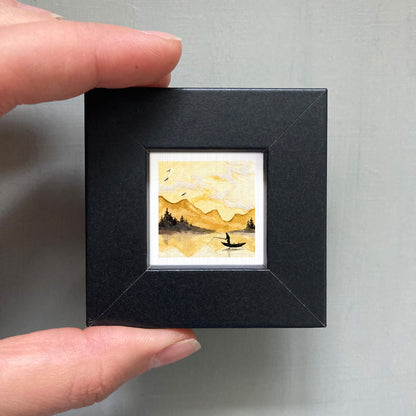 A person's hand is holding a small black frame containing a watercolor painting of a landscape with mountains and a lake.