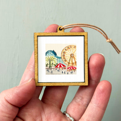 A hand holding a small wooden frame containing a picture of a carnival scene with a ferris wheel and other rides.