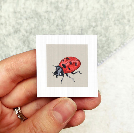 A hand holding a small square card with a watercolor illustration of a ladybug on it.