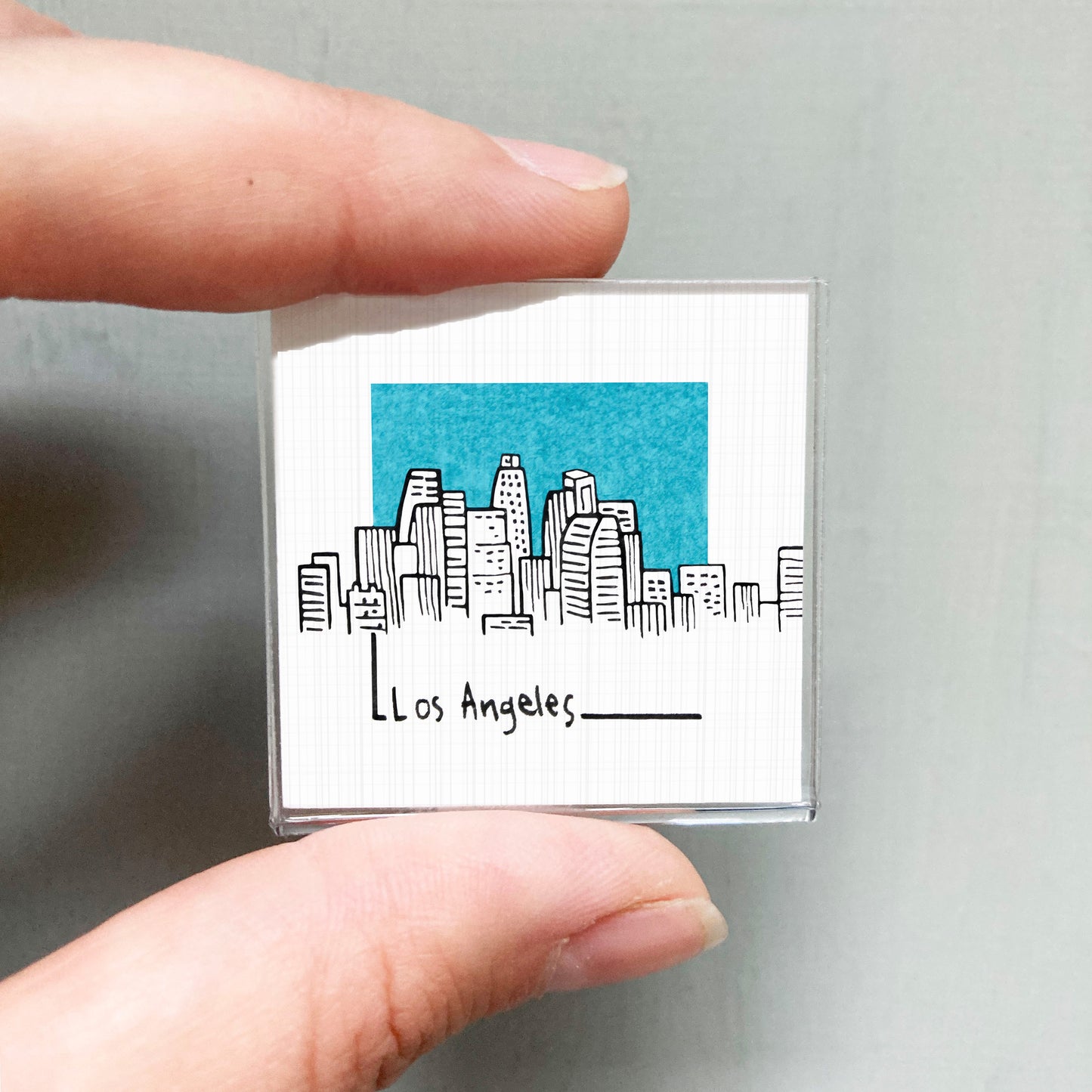 A person's hand holding a small square magnet with a drawing of a city skyline and the text "Los Angeles" written below it.
