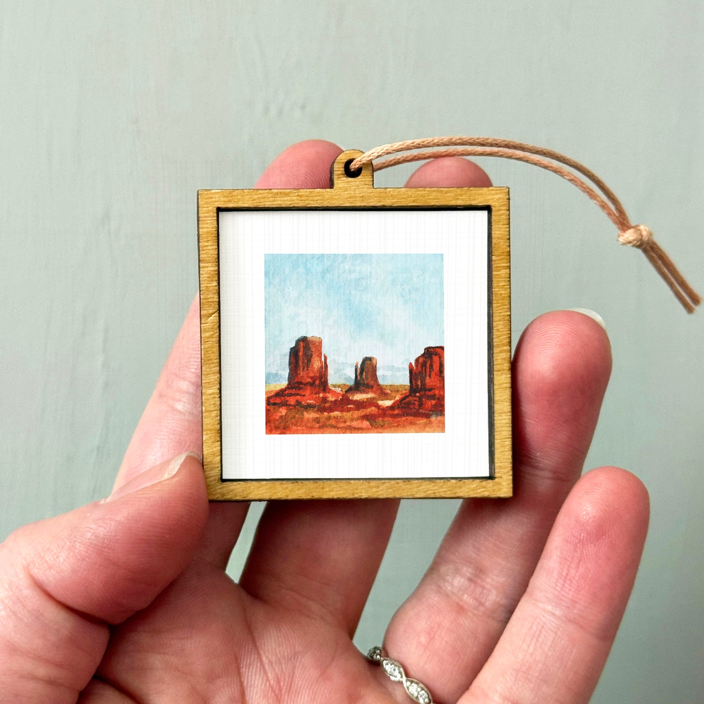 A hand holding a small wooden frame containing a painting of a desert landscape.