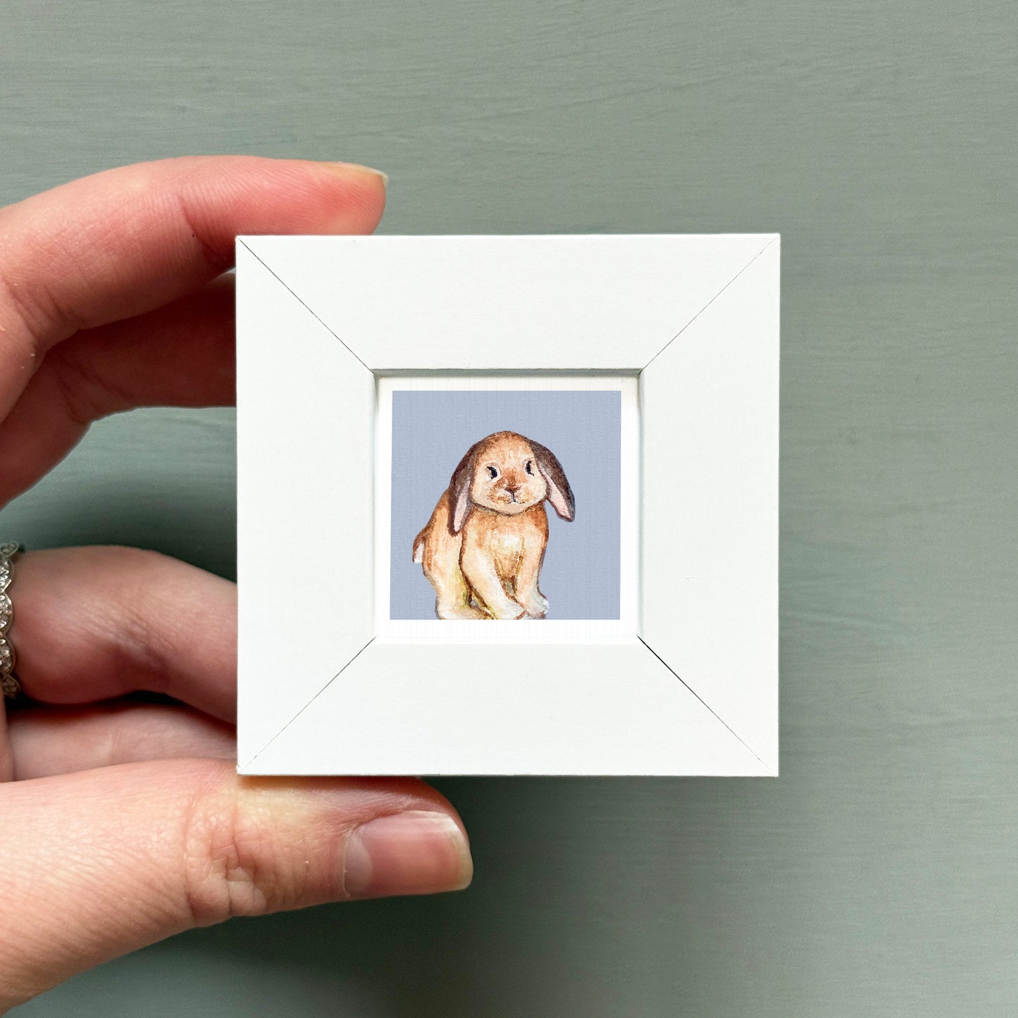 A hand is holding a small white square frame containing a watercolor painting of a rabbit.