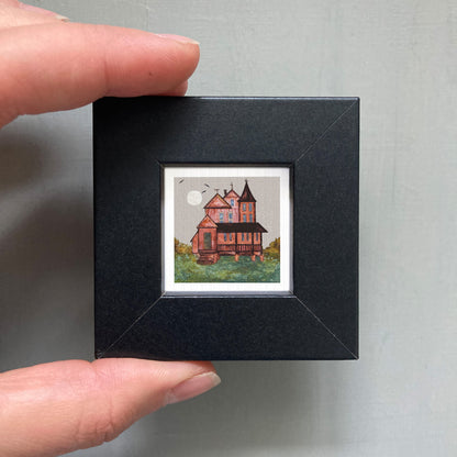 A hand is holding a small, square-shaped black frame containing a miniature painting of a house with a castle-like structure.