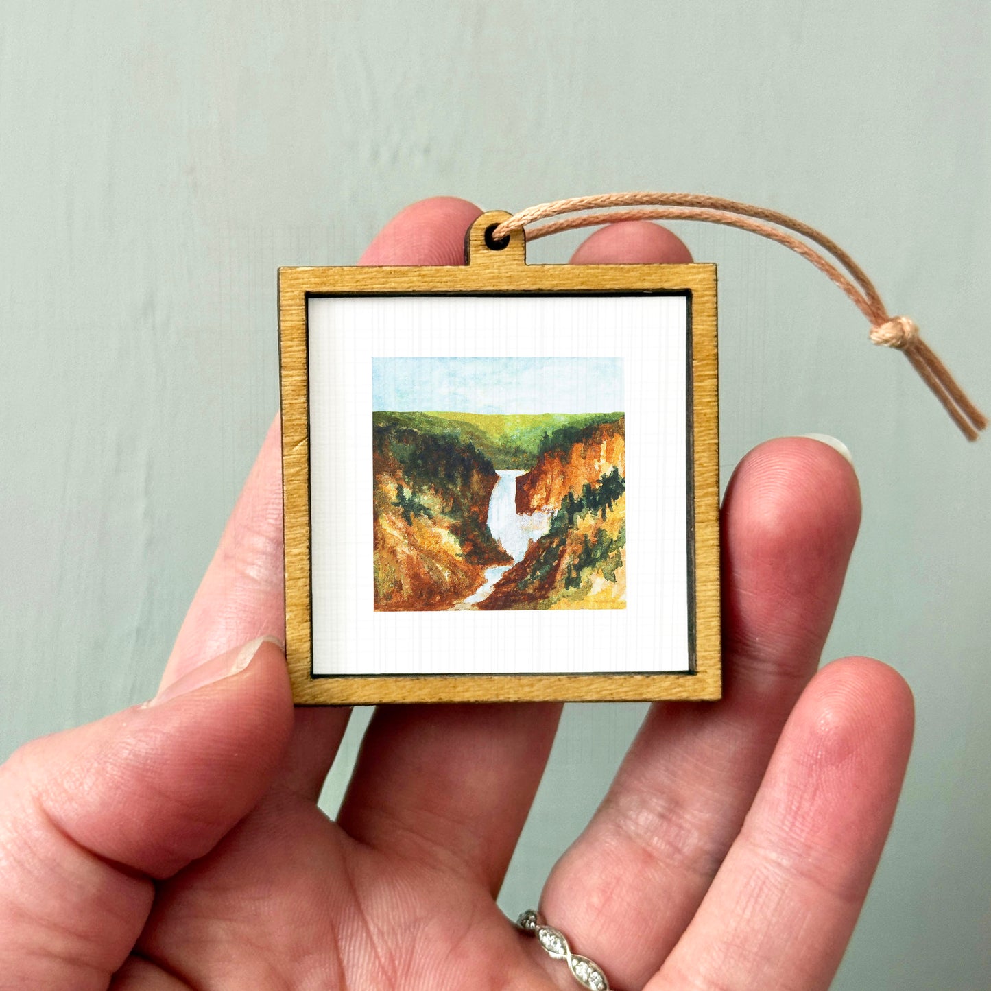 A person's hand holding a small wooden frame containing a painting of a waterfall.