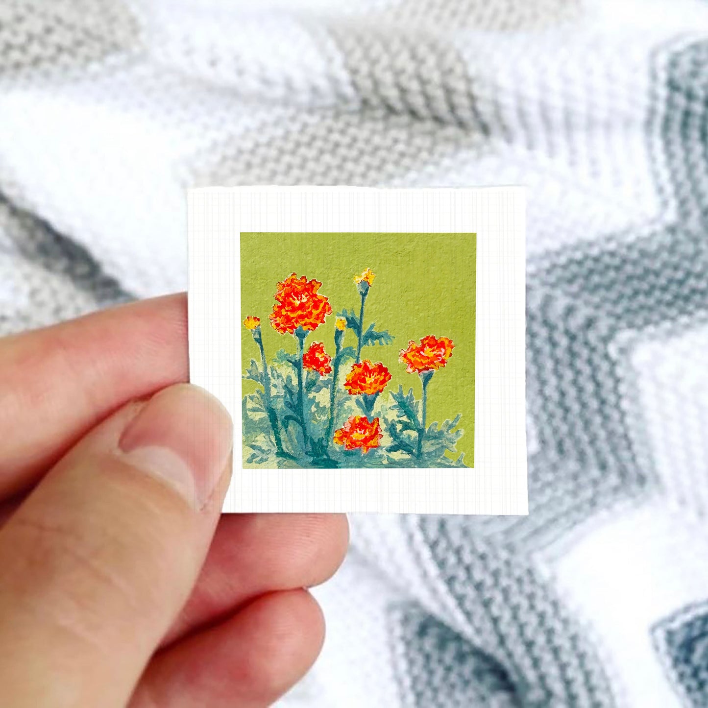 A hand is holding a small square card with a painting of red flowers against a green background.