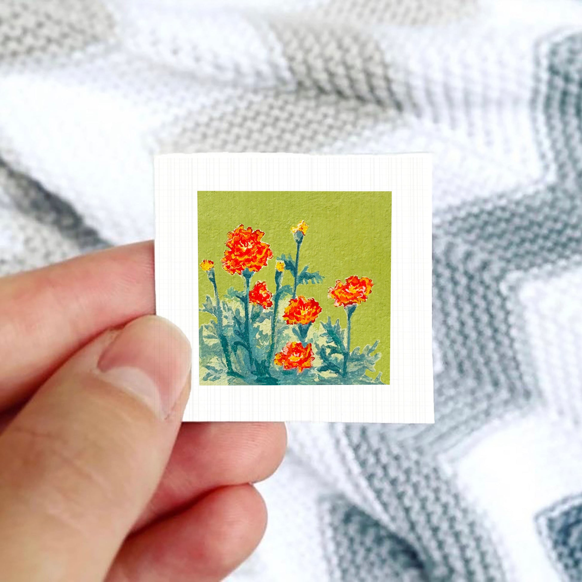 A hand is holding a small square card with a painting of red flowers against a green background.