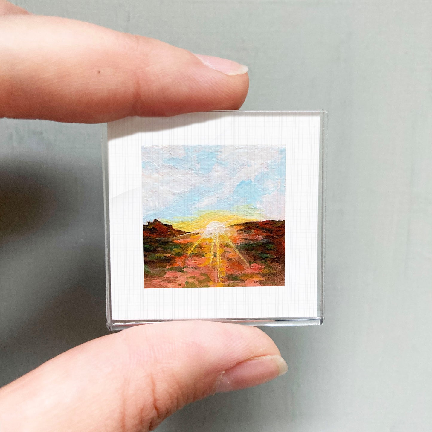 A person's hand holding a small square picture of a landscape with a sunset.